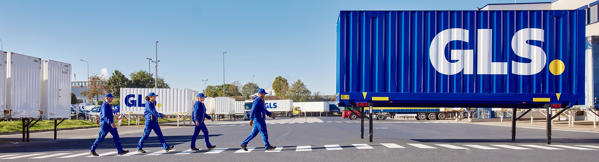 GLS team walking across a secured pedestrian crossing in front of a blue semi-trailer at a parcel logistics distribution center.